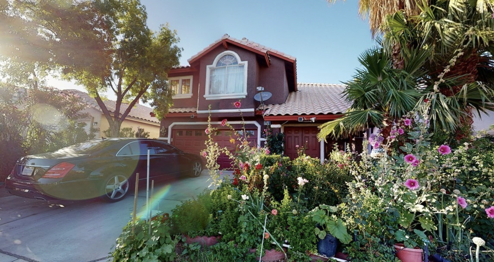 A burgundy-colored home with a lush front garden including deciduous and palm trees, roses, other flowers, and vegetables